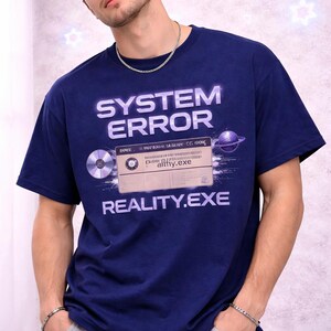 以下が含まれることがあります： 未来的なフォントで「SYSTEM ERROR」と「REALITY.EXE」の文字が入ったネイビーブルーのTシャツ。グラフィックにはカセットテープ、CD、惑星が含まれています。Tシャツは人が着ています。