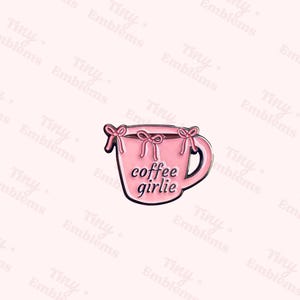 May include: A pink enamel pin in the shape of a coffee mug, featuring the words "coffee girlie" in black. The mug has a black outline and is adorned with pink bows. The background is a light pink with the words "Tiny + Emblems" repeated.