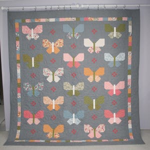 May include: A handmade quilt featuring a repeating butterfly pattern in various colors, including pink, orange, blue, and green, set against a gray background. The quilt is displayed on a wall, showcasing its intricate design and craftsmanship.