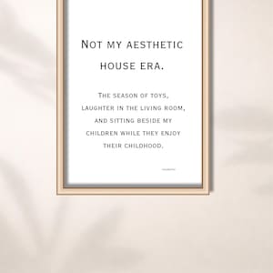Op de afbeelding: Een ingelijste print met de tekst "NOT MY AESTHETIC HOUSE ERA." De print bevat ook de tekst: "THE SEASON OF TOYS, LAUGHTER IN THE LIVING ROOM, AND SITTING BESIDE MY CHILDREN WHILE THEY ENJOY THEIR CHILDHOOD."