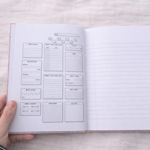 May include: Notebook with a pink cover, open to a daily planner spread. The left page has sections for goals, tasks, and water intake. The right page is lined for notes. The notebook is held open by a hand.
