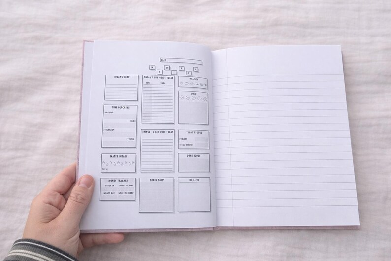 May include: Notebook with a pink cover, open to a daily planner spread. The left page has sections for goals, tasks, and water intake. The right page is lined for notes. The notebook is held open by a hand.