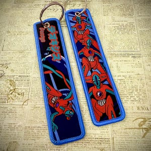 May include: Two rectangular keychains with a blue border and a silver key ring. The keychains feature a red and blue graphic design with Japanese text. The design includes a red robot-like figure.