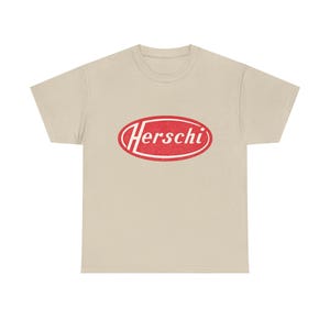 Può includere: T-shirt beige con un logo ovale rosso con la scritta "Herschi" in un font in stile vintage. La maglietta è realizzata in un materiale morbido e ha un classico girocollo. Il logo è leggermente usurato, conferendogli un aspetto retrò.