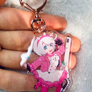 May include: A clear acrylic keychain featuring a colorful anime-style character in a pink outfit, holding a weapon. The keychain has a heart-shaped clasp in a rose gold finish.