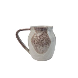 May include: A handmade ceramic mug with a white base and a brown, speckled design. The mug has a curved handle and a slightly flared rim. The brown design is concentrated in the center and around the rim.