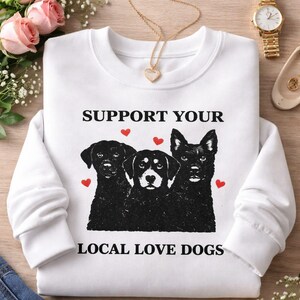 Peut inclure: Sweat-shirt blanc avec un motif de trois chiens noirs et des cœurs rouges. Le texte "SUPPORT YOUR LOCAL LOVE DOGS" est imprimé au-dessus du motif de chien. Le sweat-shirt est sur une surface en bois avec des fleurs et des bijoux.