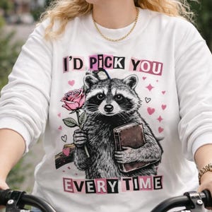 Peut inclure: Sweat-shirt blanc avec un dessin de raton laveur tenant une rose et un livre. Le texte "I'd Pick You Every Time" est imprimé en rose et noir. Le sweat-shirt est à manches longues.