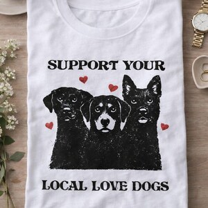 Peut inclure: T-shirt blanc avec un motif de trois chiens et le texte "SUPPORT YOUR LOCAL LOVE DOGS". Le motif comprend des illustrations de chiens noirs et des cœurs rouges. Le t-shirt est plié sur une surface en bois.