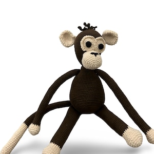 May include: A handmade crochet monkey toy in brown and cream. The monkey has long limbs, large ears, and a cream-colored face, hands, and feet. The monkey's eyes are black, and it has a small tuft of brown yarn on its head.
