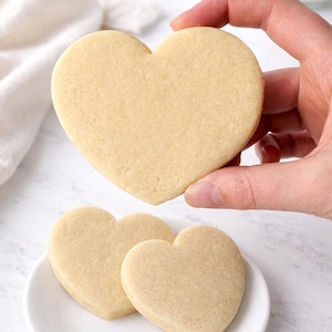 Puede incluir: Galletas de azúcar en forma de corazón. Una galleta se sostiene en alto, mientras que otras dos descansan sobre un plato blanco. Las galletas son de color marrón claro y parecen recién horneadas. Un paño blanco y un tarro de polvo blanco están en el fondo.