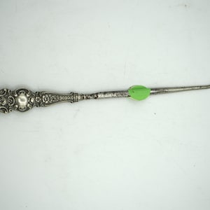 May include: A silver-toned antique crochet hook with an ornate handle and a curved hook end. The handle features detailed floral and scrollwork designs. A green sticker is affixed to the shaft. The hook is likely used for crafting.