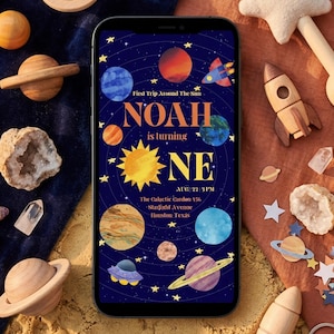 Puede incluir: Un smartphone muestra una invitación de cumpleaños con temática espacial con el texto "NOAH cumple UN año." Planetas, cohetes y estrellas de madera rodean el teléfono. La invitación incluye el texto "First Trip Around The Sun."