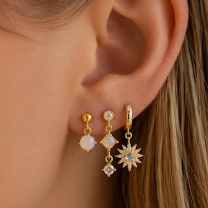 Peut inclure: Boucles d'oreilles dorées avec trois designs différents. L'une présente une petite opale ronde. Une autre a une opale en forme de losange et une pierre carrée transparente. La troisième est une créole avec un motif d'éclat d'étoile et une petite opale ronde.