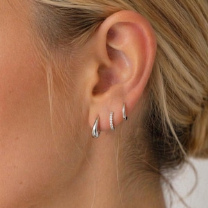 May include: Close-up of an ear with three silver earrings. The earrings include a teardrop-shaped hoop, a small hoop with embedded crystals, and a simple small hoop. The person's hair is styled in a bun.
