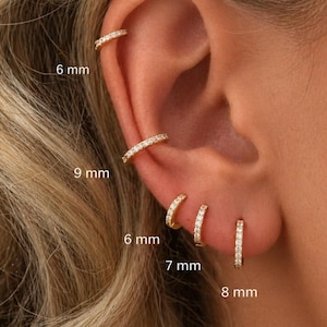 May include: A collection of gold-colored hoop earrings, each encrusted with small, clear gemstones. The earrings vary in size, with measurements of 6 mm, 7 mm, 8 mm, and 9 mm. One earring is a cuff style. The earrings are displayed on an ear.