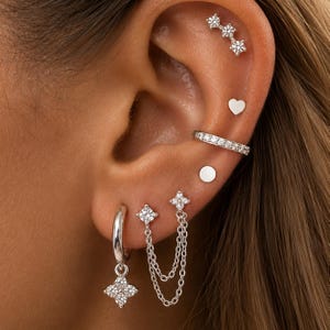 Può includere: Una collezione di orecchini color argento indossati in molteplici fori. Il set include un orecchino a tre pietre, un piccolo cuore, un cerchio con cristalli, un orecchino circolare piatto, un cerchio con ciondolo a fiore e un orecchino a doppia catena.