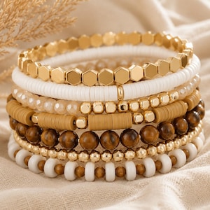 Peut inclure: Un empilement de sept bracelets composés de textures et matériaux variés, incluant des perles hexagonales dorées, des disques d'argile blancs et beiges, des perles transparentes à facettes, des pierres œil-de-tigre brunes et des perles dorées.