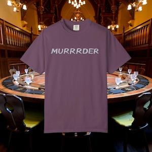 May include: A mauve t-shirt featuring the word "MURRRDER" in a silver, textured font. The shirt is the focal point, set against a blurred backdrop of a round table and chandeliers.
