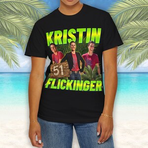 Può includere: Una t-shirt nera a maniche corte con una stampa grafica di una persona in tre pose diverse. Il testo KRISTIN FLICKINGER appare in lettere verde brillante sopra e sotto le immagini, con un cartello in legno che mostra il numero 51.