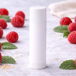 May include: A white lip balm tube stands upright, surrounded by fresh red raspberries and green mint leaves. The raspberries are scattered around the tube on a light gray surface, creating a natural and fresh aesthetic.