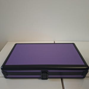 Puede incluir: Un estuche rectangular morado con una superficie texturizada y un marco negro. El estuche tiene un cierre seguro y est&aacute; dise&ntilde;ado para almacenamiento. El estuche est&aacute; sobre una superficie blanca.