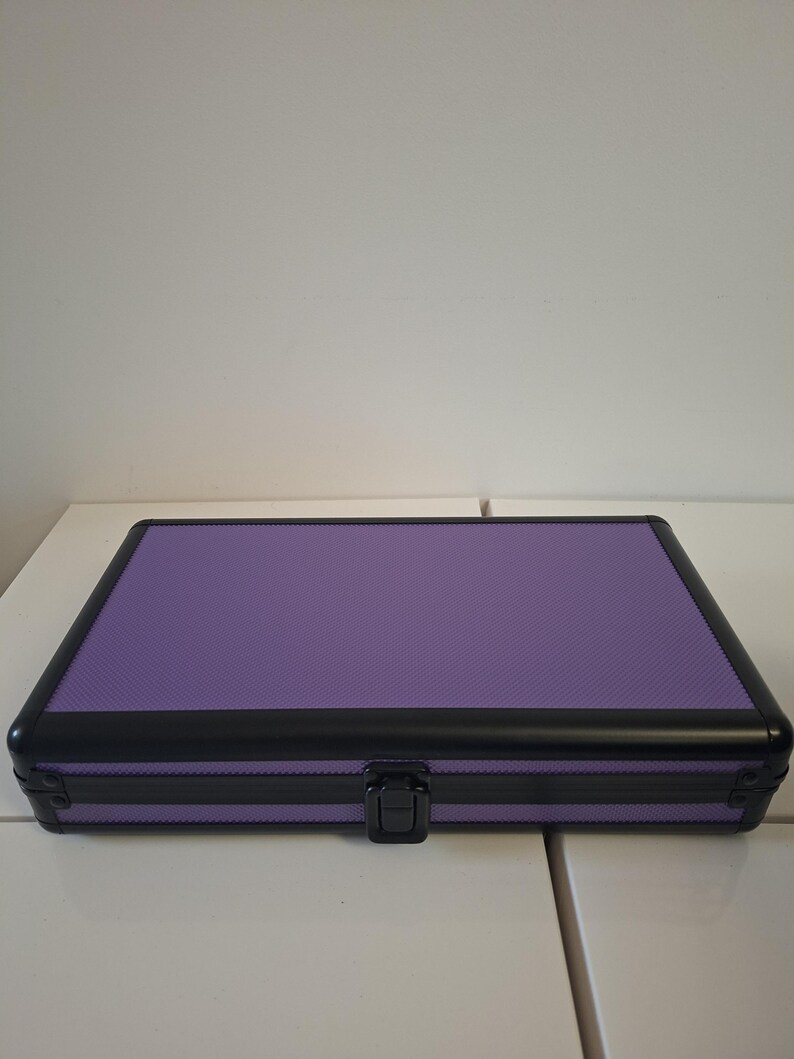 Puede incluir: Un estuche rectangular morado con una superficie texturizada y un marco negro. El estuche tiene un cierre seguro y est&aacute; dise&ntilde;ado para almacenamiento. El estuche est&aacute; sobre una superficie blanca.