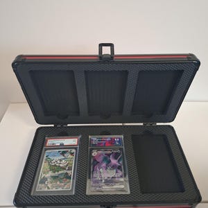 Puede incluir: Malet&iacute;n de transporte negro y rojo abierto con dos cartas coleccionables clasificadas en su interior. El malet&iacute;n tiene un asa y un interior de espuma. Las cartas est&aacute;n en fundas protectoras transparentes.