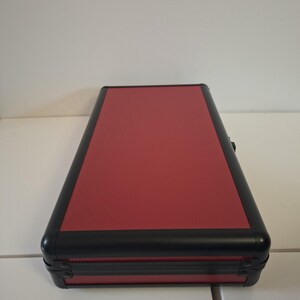 Puede incluir: Un estuche rectangular con una parte superior roja texturizada y ribete negro. El estuche tiene esquinas redondeadas y un cierre seguro. El estuche est&aacute; sobre una superficie blanca.