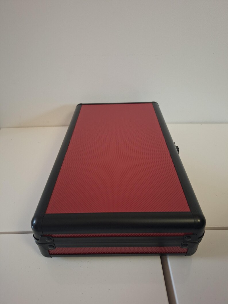 Puede incluir: Un estuche rectangular con una parte superior roja texturizada y ribete negro. El estuche tiene esquinas redondeadas y un cierre seguro. El estuche est&aacute; sobre una superficie blanca.
