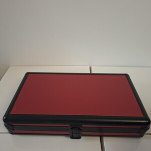Puede incluir: Un estuche rojo cerrado con marco negro y cierre negro. El estuche tiene una superficie texturizada y es de forma rectangular. El estuche est&aacute; sobre una superficie blanca.
