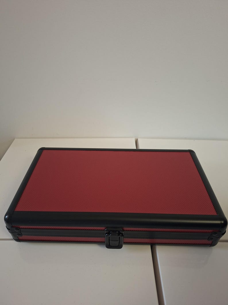 Puede incluir: Un estuche rojo cerrado con marco negro y cierre negro. El estuche tiene una superficie texturizada y es de forma rectangular. El estuche est&aacute; sobre una superficie blanca.