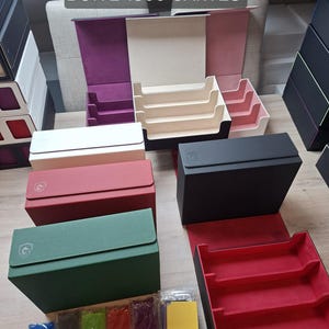May include: Various card storage boxes in red, green, black, and white, with the text "BOITE 1800 CARTES". The boxes are open and closed, revealing interior compartments. Several packs of card sleeves are also visible.