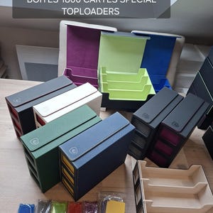 May include: Collection of card storage boxes in various colors, including green, blue, and black, with white stitching and a logo. The boxes are labeled "BOITES 1800 CARTES SPÉCIAL TOPLOADERS". Also included are various colored card sleeves.