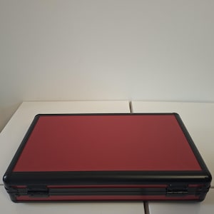 Puede incluir: Un malet&iacute;n rectangular rojo con una superficie texturizada y ribete negro. El malet&iacute;n tiene un cierre de bisagra y parece ser de metal. El malet&iacute;n est&aacute; sobre una superficie blanca.