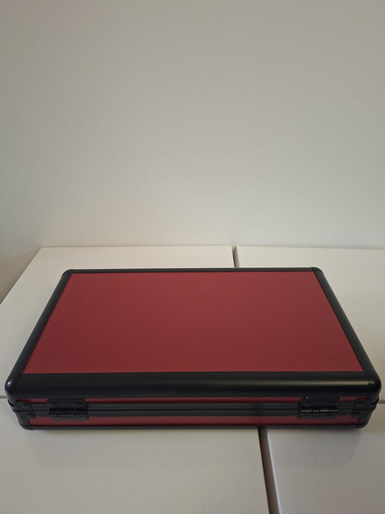 Puede incluir: Un malet&iacute;n rectangular rojo con una superficie texturizada y ribete negro. El malet&iacute;n tiene un cierre de bisagra y parece ser de metal. El malet&iacute;n est&aacute; sobre una superficie blanca.