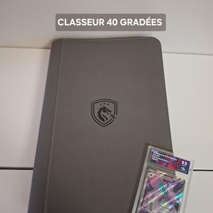 May include: A grey binder with a dragon logo and the text "CLASSEUR 40 GRADÉES". A graded trading card is next to the binder. The card is in a clear plastic case.