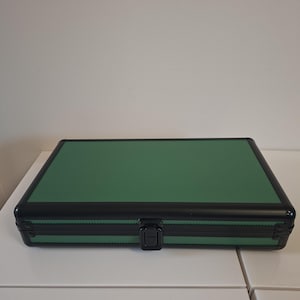 Puede incluir: Un estuche rectangular verde con una superficie texturizada y ribete negro. El estuche tiene un asa negra y un cierre de seguridad. El estuche est&aacute; sobre una superficie blanca.