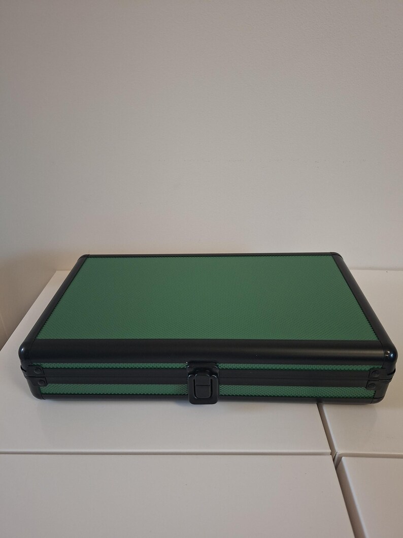 Puede incluir: Un estuche rectangular verde con una superficie texturizada y ribete negro. El estuche tiene un asa negra y un cierre de seguridad. El estuche est&aacute; sobre una superficie blanca.