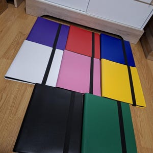 May include: A collection of eight rectangular folders in various solid colours including purple, red, blue, white, pink, yellow, black, and green. Each folder features a smooth finish and a vertical black elastic band centred to secure the cover.