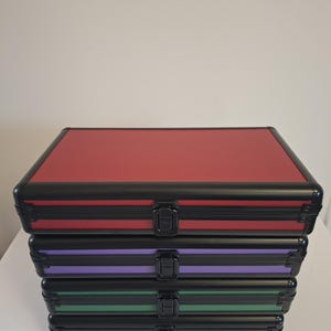 Puede incluir: Una pila de cuatro maletas de aluminio en rojo, morado, verde y negro. Cada maleta tiene un marco negro, una superficie texturizada y un cierre seguro. Ideal para almacenamiento y organizaci&oacute;n.