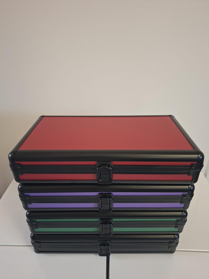 Puede incluir: Una pila de cuatro maletas de aluminio en rojo, morado, verde y negro. Cada maleta tiene un marco negro, una superficie texturizada y un cierre seguro. Ideal para almacenamiento y organizaci&oacute;n.