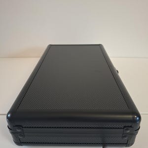 Puede incluir: Un estuche rectangular negro con una superficie texturizada y esquinas redondeadas. El estuche tiene un marco de metal y est&aacute; cerrado. El estuche est&aacute; sobre una superficie blanca.