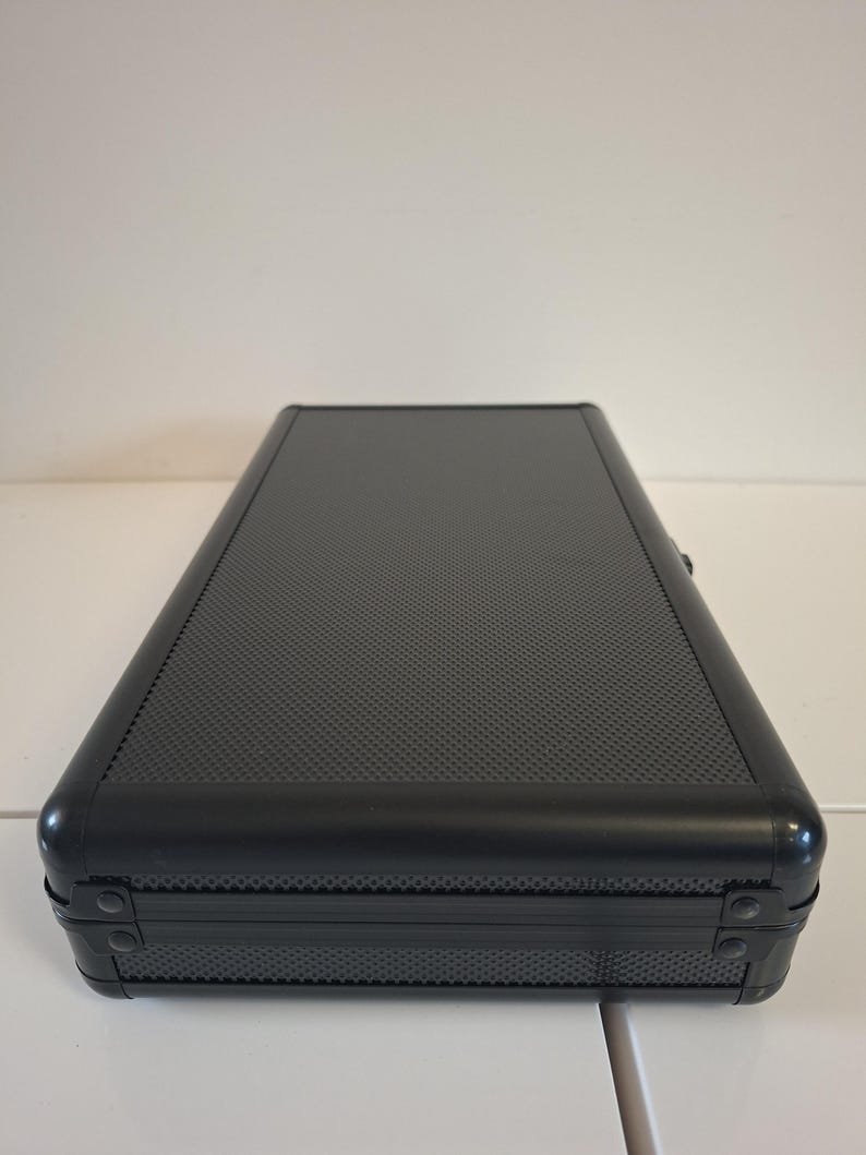 Puede incluir: Un estuche rectangular negro con una superficie texturizada y esquinas redondeadas. El estuche tiene un marco de metal y est&aacute; cerrado. El estuche est&aacute; sobre una superficie blanca.