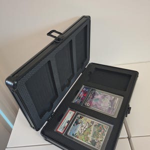 Puede incluir: Malet&iacute;n de aluminio negro abierto con dos cartas coleccionables clasificadas. El malet&iacute;n tiene un interior texturizado y un asa. Las cartas est&aacute;n en fundas protectoras.