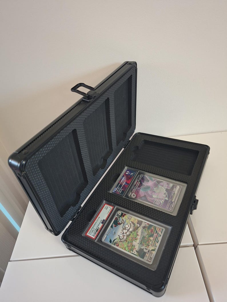 Puede incluir: Malet&iacute;n de aluminio negro abierto con dos cartas coleccionables clasificadas. El malet&iacute;n tiene un interior texturizado y un asa. Las cartas est&aacute;n en fundas protectoras.