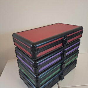 Puede incluir: Una pila de cuatro maletas de aluminio negro con detalles de color. La maleta superior es roja, seguida de morada, verde y negra. Cada maleta tiene un asa negra y un cierre plateado. Las maletas est&aacute;n apiladas.