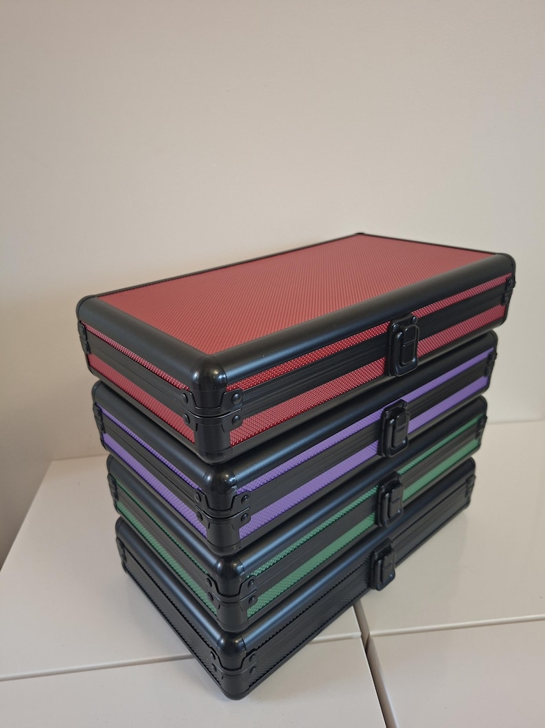 Puede incluir: Una pila de cuatro maletas de aluminio negro con detalles de color. La maleta superior es roja, seguida de morada, verde y negra. Cada maleta tiene un asa negra y un cierre plateado. Las maletas est&aacute;n apiladas.