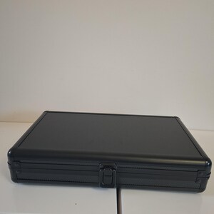 Puede incluir: Un estuche rectangular negro con una superficie texturizada y un cierre central. El estuche est&aacute; cerrado y parece ser de metal. El estuche est&aacute; sobre una superficie blanca.