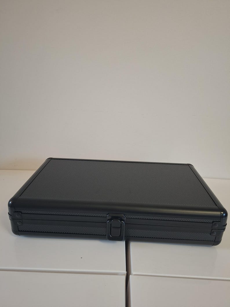 Puede incluir: Un estuche rectangular negro con una superficie texturizada y un cierre central. El estuche est&aacute; cerrado y parece ser de metal. El estuche est&aacute; sobre una superficie blanca.